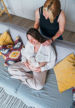 Photo for Young woman tender massaging pregnant partner's female shoulders. Same-sex marriage couple on home living room sofa. Woman's health, happy pregnancy doula supporting and calm mental mood concept image - Royalty Free Image