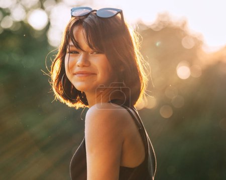 Photo for A beautiful young teenage girl dressed in a black dress cheerfully smiling at the camera with a warm sunset backlight. Beautiful people and a fashion concept image. - Royalty Free Image
