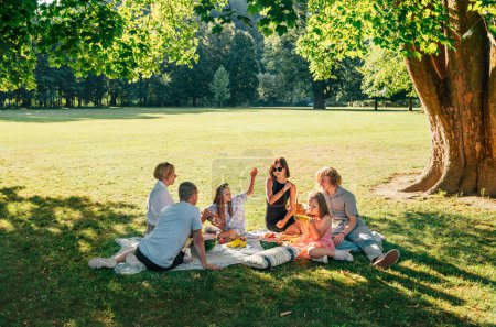 Photo for Big family sitting on picnic blanket in city park linden treeb during weekend Sunday sunny day. They are chatting and eating boiled corn and watermelon. Family values and outdoors activities concept - Royalty Free Image