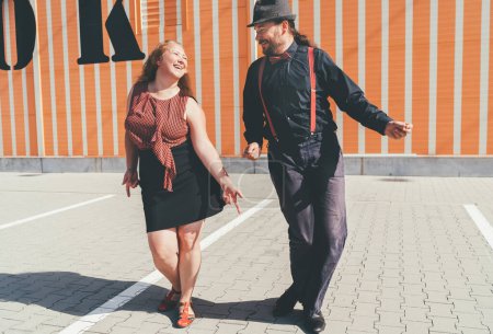 Fröhliches Paar, das einen Retro-Swing-Tanz zu Jazzmusik tanzt, der während der West Coast Swing Ära in den 1920er-40er Jahren sehr populär war. Sie lächeln einander neben gestreiftem Wandhintergrund zu.
