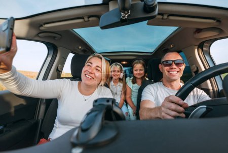 Photo for Happy young couple with two daughters inside car during auto trop. They are smiling, laughing and taking selfies using smartphone. Family values, traveling, social media and new technology concepts. - Royalty Free Image