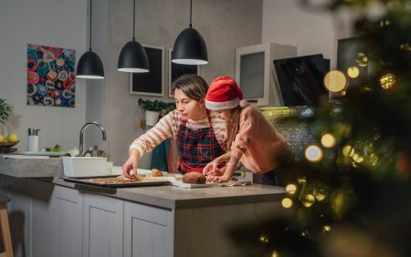 Foto de Linda niña en el sombrero rojo de Santa con la madre haciendo galletas caseras de jengibre de Navidad usando cortadores de galletas juntos en la cocina casera. Felices vacaciones en familia preparación y concepto de infancia - Imagen libre de derechos