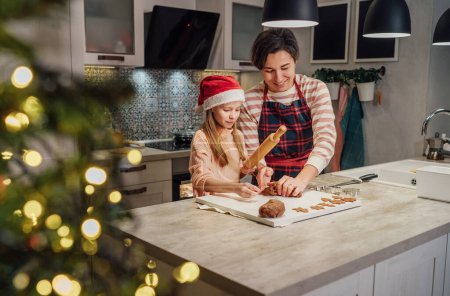 Foto de Cute little girl in red Santa hat with mother making homemade Christmas gingerbread cookies using rolling pin together in home kitchen. Happy family holidays preparation and childhood concept. - Imagen libre de derechos