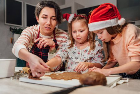 Photo for Cute little girls in red Santa hats with mother making homemade dough Christmas gingerbread cookies using cookie cutters together in home kitchen. Happy family holidays preparation, childhood concept - Royalty Free Image