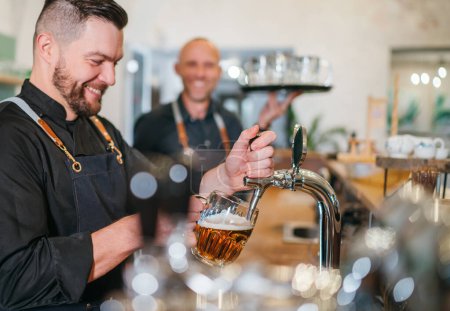 Photo for Stylish bearded barman dressed black uniform beer tapping at bar counter and waiter with tray chatting smiling to each other.Successful people team work friendship and good relationships concept image - Royalty Free Image