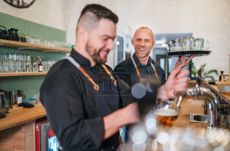 Photo for Two men friends barman and waiter with tray dressed black uniform at bar counter chatting smiling to each other. Sincere people relationships, team work, men friendship and good mood concept image. - Royalty Free Image