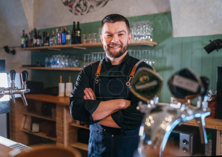 Photo for Portrait of happy smiling bearded barman dressed in a black uniform with an apron at bar counter with draught beer taps. Successful people, hard work, consumer cafes and restaurants industry concept. - Royalty Free Image
