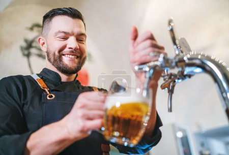 Photo for Smiling stylish bearded barman dressed black uniform with an apron tapping fresh lager beer into pilsner glass mug at bar counter. Successful people, beer consumption, beverages industry concept image - Royalty Free Image