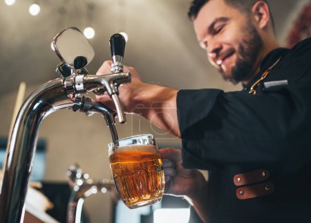 Photo for Tapping fresh lager beer in glass mug close up. Smiling stylish bearded barman dressed black uniform with an apron at bar counter. Successful people, beer consumption, beverages industry concept imag - Royalty Free Image