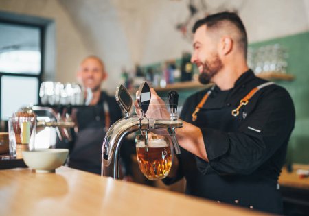 Photo for Fresh lager beer glass mug and barman dressed uniform smiling to teammate waiter with tray beer tapping at bar counter. Successful people teamwork, friendship, brewing, restaurant industry concept - Royalty Free Image