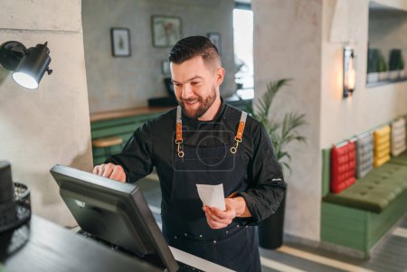 Stylish bearded smiling waiter dressed black uniform processing customer orders using point of sale order terminal system touch screen. Successful people teamwork, restaurant industry concept image.