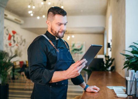 Smiling small business owner dressed in a black chef uniform with an apron using digital tablet in his cozy restaurant hall. Successful people, hard work, consumer cafes, restaurants industry concept.