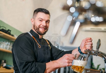 Photo for Smiling stylish bearded barman dressed uniform with apron gazing at camera while tapping fresh lager beer glass mug at bar counter.Successful people, beer consumption, beverages industry concept image - Royalty Free Image