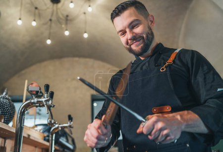Photo for Smiling bearded chef cook dressed black uniform with apron sharpening knife with Honing Steel tool. Professional occupation, hard work, food preparation, restaurants industry low angle view shot. - Royalty Free Image