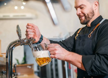 Sonriente barman barbudo elegante vestido uniforme negro con un delantal golpeando cerveza cerveza cerveza fresca en taza de vidrio en el mostrador del bar. Personas exitosas, consumo de cerveza, bebidas imagen conceptual de la industria