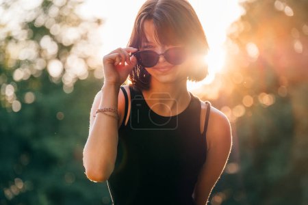 Photo for Beautiful young teenage girl in fancy sunglasses dressed in black dress challenging smiling at the camera with a warm sunset backlight. Beautiful people and a fashion concept image. - Royalty Free Image