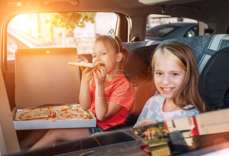 Téléchargez les photos : Deux sœurs souriantes positives sont heureuses de manger des pizzas italiennes juste cuites assises dans des sièges auto pour enfants sur le siège arrière. Bonne enfance, restauration rapide, ou voyage auto pause déjeuner concept image. - en image libre de droit