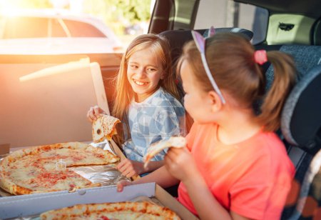 Photo for Portrait of two positive smiling sisters eating just cooked italian pizza sitting in child car seats on car back seat. Happy childhood, fastfood eating or auto jorney lunch break concept image. - Royalty Free Image
