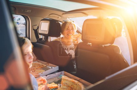 Téléchargez les photos : Portrait de jeune femme mangeant juste cuit pizza italienne alors qu'elle voyageait en famille dans une voiture moderne. Joyeux moments et valeurs de la famille, restauration rapide ou voyage auto pause déjeuner concept image. - en image libre de droit