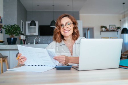 Beautiful middle-aged woman in glasses smiling at camera. She does Quarterly financial reports at the home office using modern laptop. Small business, home finance, money savings concept image.