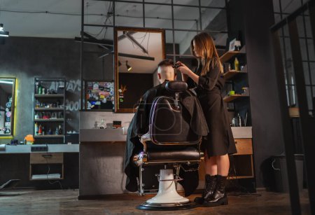 Foto de Hermoso peluquero femenino joven que hace el peinado de los hombres usando Haircut Clipper en el interior de la barbería de estilo loft moderno. Servicios de cuidado del cabello pequeñas empresas locales, cosméticos e industria del cuidado personal. - Imagen libre de derechos