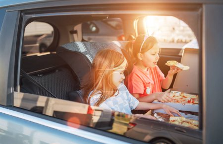 Téléchargez les photos : Deux petites sœurs sont heureuses de manger des pizzas italiennes cuites sur le siège arrière de la voiture. Bonne enfance, restauration rapide, ou voyage auto pause déjeuner concept image. - en image libre de droit