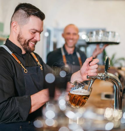 Stylish bearded barman dressed black uniform beer tapping at bar counter and waiter with tray chatting smiling to each other.Successful people team work friendship and good relationships concept image