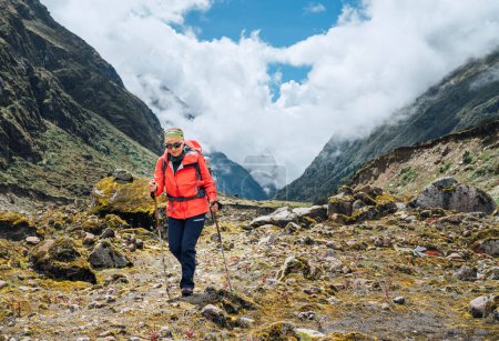 Woman in sunglasses with backpack and trekking poles dressed red softshell jacket hiking during Makalu Barun National Park trek in Nepal. Mountain hiking, traveling and active people concept image.