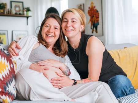 Happy smiling women couple waiting for baby. Same-sex pregnant marriage couple on home living room sofa. Woman's health, happy pregnancy doula supporting and calm mental mood concept image