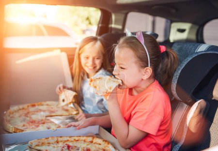 Téléchargez les photos : Portrait de deux sœurs souriantes positives mangeant des pizzas italiennes juste cuites assises dans des sièges auto pour enfants sur le siège arrière de la voiture. Bonne enfance, jeûne manger ou auto jorney pause déjeuner concept image. - en image libre de droit