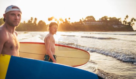 Téléchargez les photos : Adolescent avec une planche de surf va à la mer surf. Il passe des vacances d'hiver et profite d'un magnifique coucher de soleil avec son père sur l'île de Sri Lanka. Concept de vacances actives familiales. - en image libre de droit