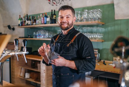 Porträt eines glücklich lächelnden, bärtigen Barmanns in schwarzer Uniform mit Schürze, der das Bierglas an der Theke wischt. Erfolgreiche Menschen, harte Arbeit, Verbrauchercafés und Restaurantkonzept.