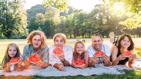 Große Familie auf der Picknickdecke im Stadtpark unter der Linde während des Sonntags am Wochenende, lächelt, lacht in die Kamera und isst Wassermelone. Familienwerte und Outdoor-Konzept.