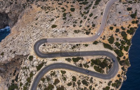 Einsamer weißer Kleinwagen, der durch die Serpentinenkurve einer asphaltierten Bergstraße in der Nähe des Leuchtturms von Cap de Formentor mit Meereslandschaft und felsiger Küste fährt. Mallorca, Balearen, Spanien. Luftaufnahme von oben.