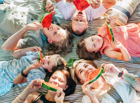 Kleine Töchter mit der Familie der Eltern liegen am sonnigen Wochenende auf einer Picknickdecke, lächeln, lachen und essen rote saftige Wassermelonenstücke. Familienwerte, Obst-Vitamine, Outdoor-Zeitkonzept