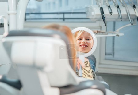 Photo for Little girl sitting in stomatology clinic chair and smiling at mirror showing her teeth after teeth dental procedures. Healthcare, kid's health and medicare industry concept image - Royalty Free Image