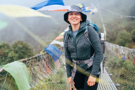 Young smiling backpacker woman crossing canyon over Suspension Bridge with multicolored Tibetan Prayer flags. Mera peak climbing route trek Lukla Sagarmatha National Park, Nepal. Active people concept