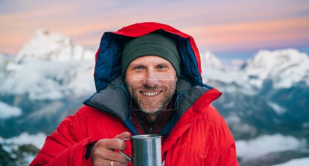 Téléchargez les photos : Portrait de sourire à la caméra alpiniste de haute altitude veste aube chaude rouge veste tenant tasse de thé chaud en métal avec fond panorama montagnes. Mera Peak High Camp 5700m, Himalaya, Népal. - en image libre de droit