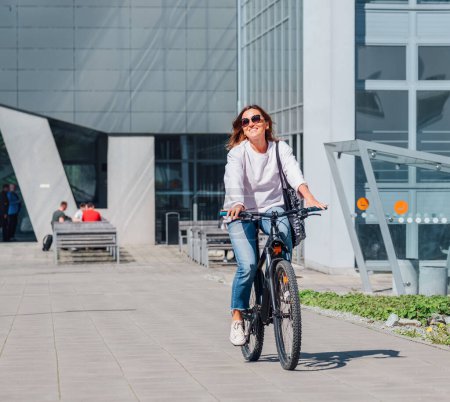 Eine glücklich lächelnde kaukasische Frau mit schicker Sonnenbrille auf einem Fahrrad auf der modernen Straße der Stadt. Ökologisch nachhaltiger Verkehr und glückliche Menschen.