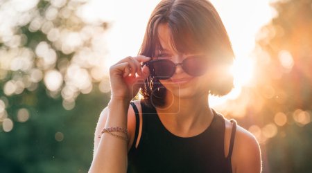 Beautiful young teenage girl in fancy sunglasses dressed in black dress challenging smiling at the camera with a warm sunset backlight. Beautiful people and a fashion concept image.
