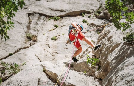 Hombre de mediana edad escalador activo en casco protector mientras rappel de la pared de roca acantilado utilizando cuerdas con un dispositivo de bajada y arnés de escalada. Concepto activo de gasto de tiempo en deportes extremos