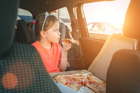 Téléchargez les photos : Portrait de petite fille de 5 ans mangeant juste cuit pizza italienne assis dans le siège auto enfant sur le siège arrière de la voiture et regardant par la fenêtre. Bonne enfance, restauration rapide ou voyage auto pause déjeuner image - en image libre de droit