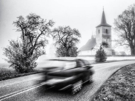 Photo for Blurred car motion on the road with old church at background. Village Ludrova in Slovakia. Black and white photo - Royalty Free Image