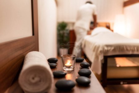Towel, candle and lava rocks in spa salon. Masseur with client lying on massage table in the background