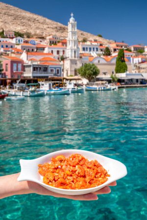 Foto de Halki, Grecia - 6 de julio de 2022: Placa con camarones rojos y mar azul en la isla de Halki al fondo - Imagen libre de derechos