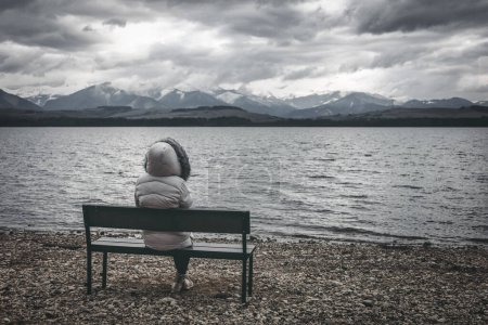 Mujer con chaqueta sentada en el banco en la orilla del lago. Cielo nublado oscuro con montañas al fondo