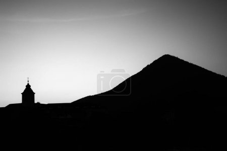 Silhouette of tower of church with cross on top and big hill at background.