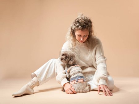 Nettes Mädchen mit Hund im Pullover auf beigem Hintergrund. Lockige Frau mit Schokopudel. gemütlich mit einem Haustier