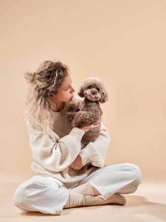 Linda chica con un perro en un suéter sobre un fondo beige. Mujer rizada con caniche de chocolate. acogedor con una mascota