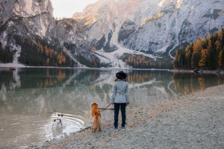 Eine Person und ihr goldener Begleiter verweilen am Ufer eines Kieselsees und blicken auf das ruhige alpine Wasser, das den Schein einer herbstlichen Dämmerung widerspiegelt.